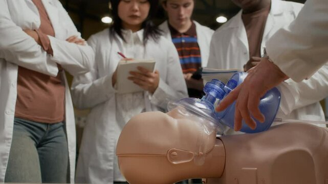 Rack focus from group of young medical students on hands of instructor teaching teenagers cardiopulmonary resuscitation with use of bag valve mask while demonstrating first aid on manikin at college