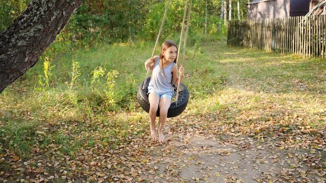 Girl sits on tire swing, happily swinging back and forth among surrounding trees. Cute girl plays outside, enjoying country vacation