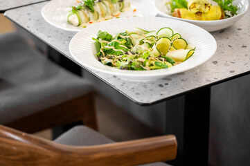 Fresh salad with cucumber slices, greens, and dressing on a white plate in the foreground, surrounded by sushi rolls and a yellow wrap dish on a wooden table.