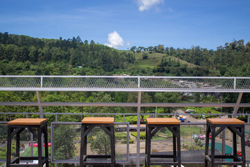 Four modern wooden bar stools line the terrace with white railings, overlooking a stunning green landscape view with trees, hills and bright blue skies.