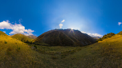 Sunlit mountain valley with grassy slopes under a bright blue sky with scattered clouds.