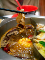 human hand using chopsticks hold meat over Sichuan mala hotpot shabu on restaurant table.