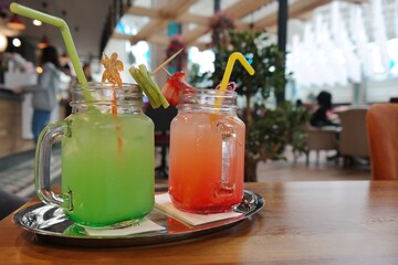 Two mason jars filled with colorful drinks, one green and one pink, garnished with straws and fruit, sit on a tray in a cozy café setting.