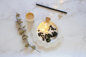 A cozy setup with a light bulb, coffee beans, a cup, a pen and eucalyptus leaves on a lace doily, creating a warm and inviting atmosphere