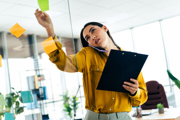 Young businesswoman multitasking in a modern office environment with a clipboard and a phone,...