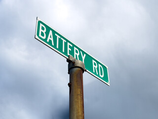 A green sign with white text, Battery Road, in raised letters on a metal plate. The location sign is on a round stainless steel post outside. The street sign is for a historic road and landmark