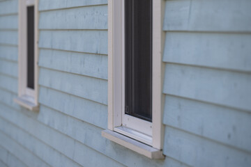 The exterior wall of a pale blue vintage house has two glass, white vinyl casement windows. The cottage wall is textured horizontal wooden Cape Cod siding boards. The glass windows are closed.