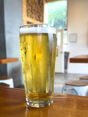 A clear pint glass filled with a pale ale. The traditional malty and earthy beer sits on a golden wooden bar table. There's a shelf in the background filled with wood junk for an indoor fireplace. 