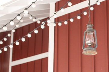 The exterior wall of a red wooden barn with white trim. There are strings of white solar lights hanging from the outside roof of the building. An antique rusty glass hurricane lamp hangs from a chain.