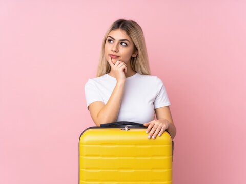 Young Uruguayan blonde woman with a suitcase full of clothes over isolated pink wall and thinking