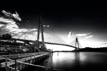 Black and white photo of a bridge over the sea at sunset.
