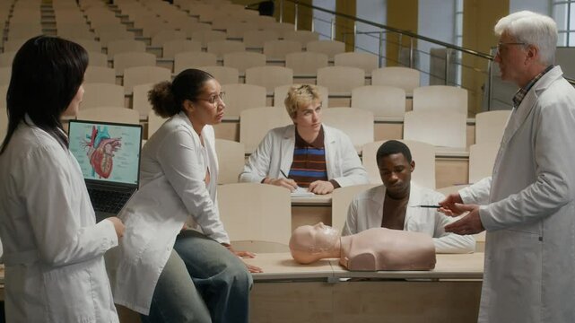 Senior male professor instructing group of young medical students in first aid in event of heart attack pointing at CPR manikin during training class in university auditorium
