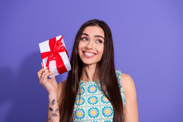 Smiling young woman holding a gift box with red ribbon in front of violet backdrop, ideal for...