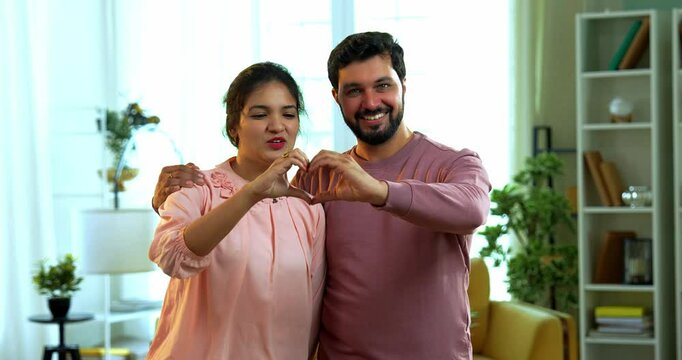 Indian Asian young couple smiling cheerfully while forming a heart shape using their hand palms together, standing in a cozy modern living room, both looking directly at camera with love and warmth