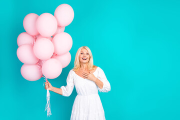 Cheerful mature woman in white dress holding pink balloons against turquoise background, celebrating with a joyful expression