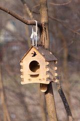 Hanging on a tree branch in a forest park in spring in sunny weather is a handmade wooden bird feeder made of plywood in the form of a little house with bird-shaped windows in the attic and a roof.