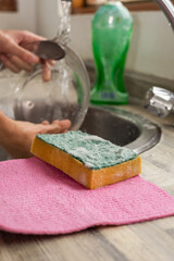 A sponge rests on a pink cloth near the sink, while someone rinses dishes in the background of a clean and bright kitchen.

