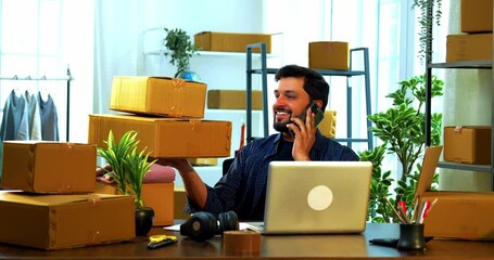 Indian Asian small businessman sitting in a warehouse store with cardboard shipment boxes around, holding a package in one hand and smiling while talking happily on smartphone confirming online order