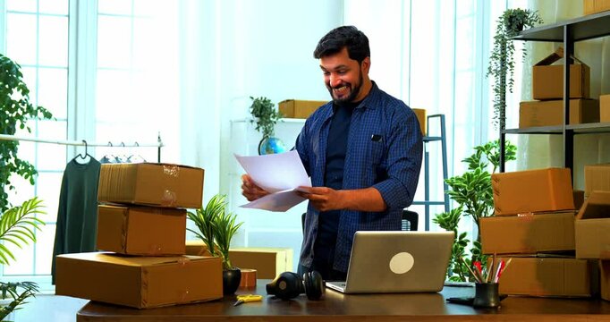 Indian Asian small businessman holding order list celebrating success with joyful expression after receiving large number of online product orders or shipment confirmation standing at store room