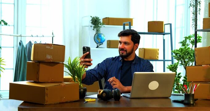 Indian Asian small businessman scanning barcode label on cardboard carton box using smartphone camera while sitting at desk in store room office, working on online order or shipment processing task