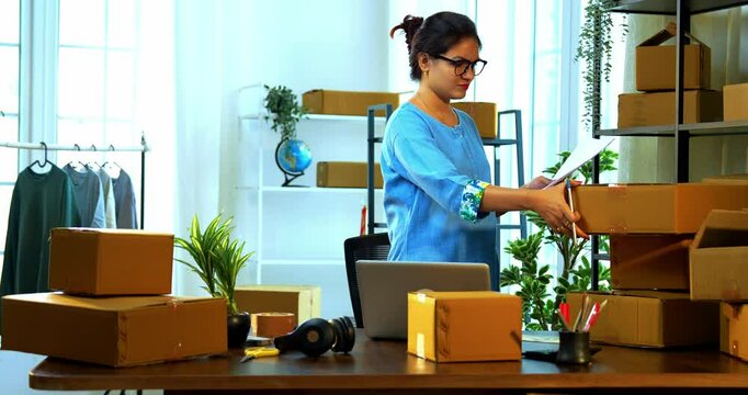 Indian Asian small businesswoman checking and maintaining checklist of shipment boxes in warehouse office, carefully updating inventory records and ensuring packaging accuracy for order dispatch