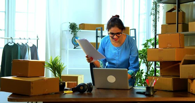 Indian Asian small businesswoman working on laptop while sitting at desk in a store room filled with cardboard boxes, entering shipment details from printed documents for order dispatch processing