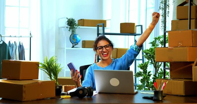 Indian Asian small businesswoman using smartphone and suddenly celebrating success with happy expressions, receiving good news or online order confirmation, feeling joyful sitting in store office desk