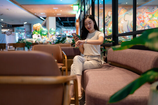 Young Asian businesswoman checking time on smartwatch while using smartphone in shopping mall cafe