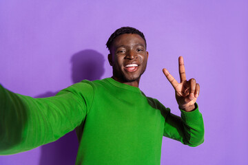 Young man in green sweatshirt showing peace gesture against a purple background