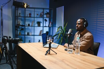 Black vlogger filming podcast on smartphone in studio. Cheerful blogger