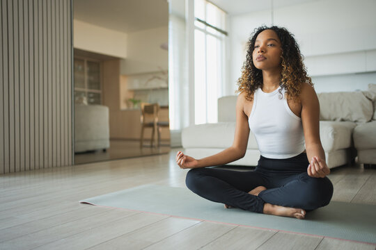 Yoga mindfulness meditation. Young healthy African girl practicing yoga at home. Woman sitting in lotus pose on yoga mat meditating relaxing indoor. Girl doing breathing practice. Yoga at home