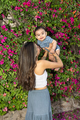 Mother and son playing, mother holding a baby pointing to the sky, the baby is smiling and in the background there is a wall with plants, and both are wearing a cool outfit