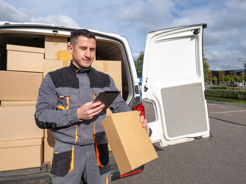 Delivery worker in uniform stands at the back of a van full of packages, scanning a parcel with a tablet during a delivery route on a sunny day.