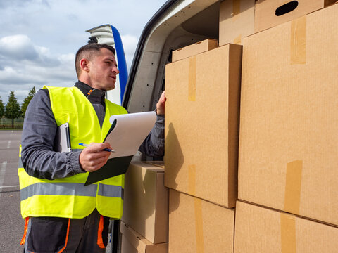 Delivery worker in a reflective vest checks a clipboard while organizing stacked cardboard boxes in the back of a van during a delivery or logistics operation.