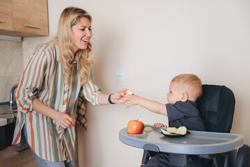 Mother giving apple to her little son sitting in high chair
