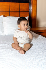 Baby sitting in bed looking to the side with finger in mouth and wearing white clothes