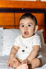 Baby sitting in bed with a surprised face, opening his mouth and eyes and wearing white clothes