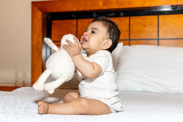 Baby sitting on a bed holding a stuffed animal and the baby has a frustrated face
