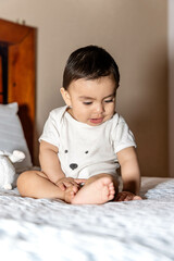 Baby sitting on a bed, touching the bed with his hand and looking surprised, and next to him there is a stuffed animal and a pillow
