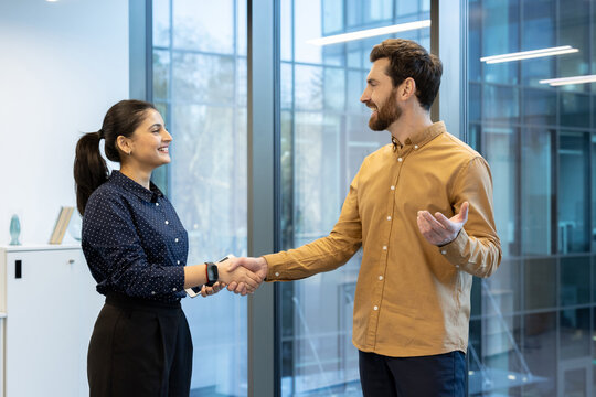 A man and a woman shaking hands in an office, symbolizing agreement or partnership. The scene is bright with natural lighting, suggesting a modern workplace.