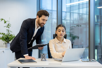 A business meeting is observed as a man and a woman collaborate in a modern office setting. She works on a laptop while he holds a clipboard.