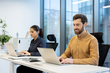 Two colleagues work collaboratively in a bright office, each using a laptop with a focused expression, showcasing teamwork.