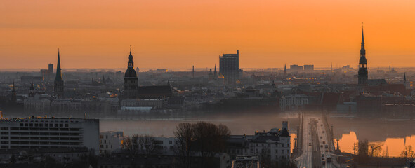A warm sunrise illuminates Riga's old town, featuring St. Peter's Church, Riga Cathedral, a bridge...