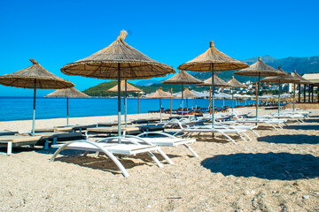 Straw beach umbrellas and comfortable sun loungers on clean sand and pebble beach. Himare. Albania.