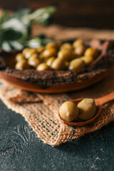 Rustic wooden bowl filled with green olives on burlap fabric with olive branch in background....