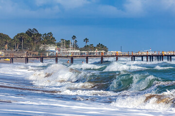 Turquoise waves crash on Sorgun beach as two people swim under the sun. Palms and a pier with umbrellas in the background. Side, Turkey, Mediterranean.