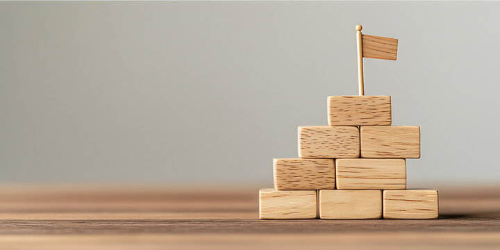 Wooden block tower with flag represents goals and success, achieved step by step. Strategy and planning are key for accomplishment.