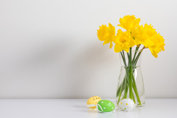Easter background with yellow flowers in a glass vase and colored Easter eggs against a white wall, front view.