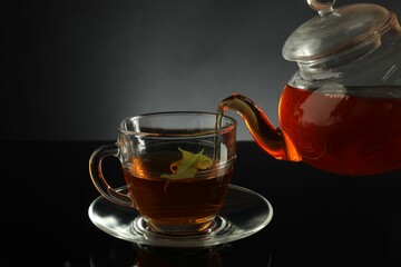 Pouring freshly brewed tea from teapot into glass cup on black table, closeup