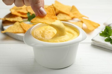 Woman dipping nacho chip into tasty cheese sauce at white wooden table, closeup